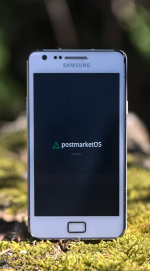 A Samsung Galaxy S II showing the postmarketOS boot splash sitting on moss in nature