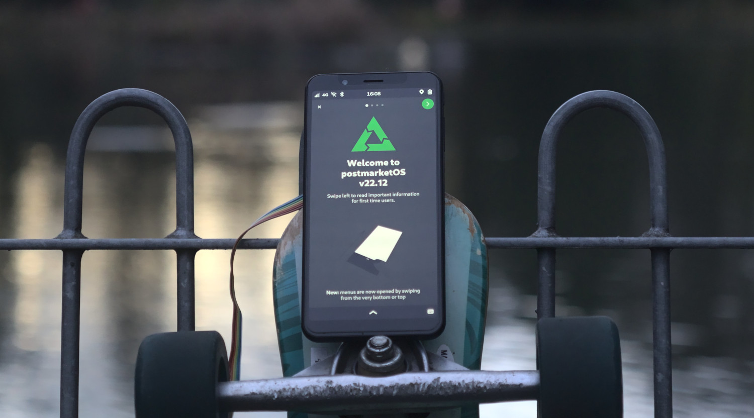 A skadeboard leaning on a railing infront of a river, with a smartphone on top of one axe displaying the welcome app of postmarketOS v22.12