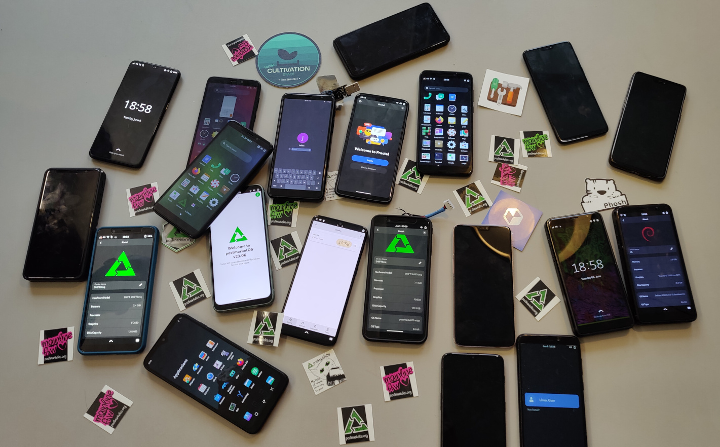 A lot of phones on a table, most of them running postmarketOS (also one with a Debian logo), and lots of stickers, mostly for postmarketOS, and a beer coaster of the cultivation space